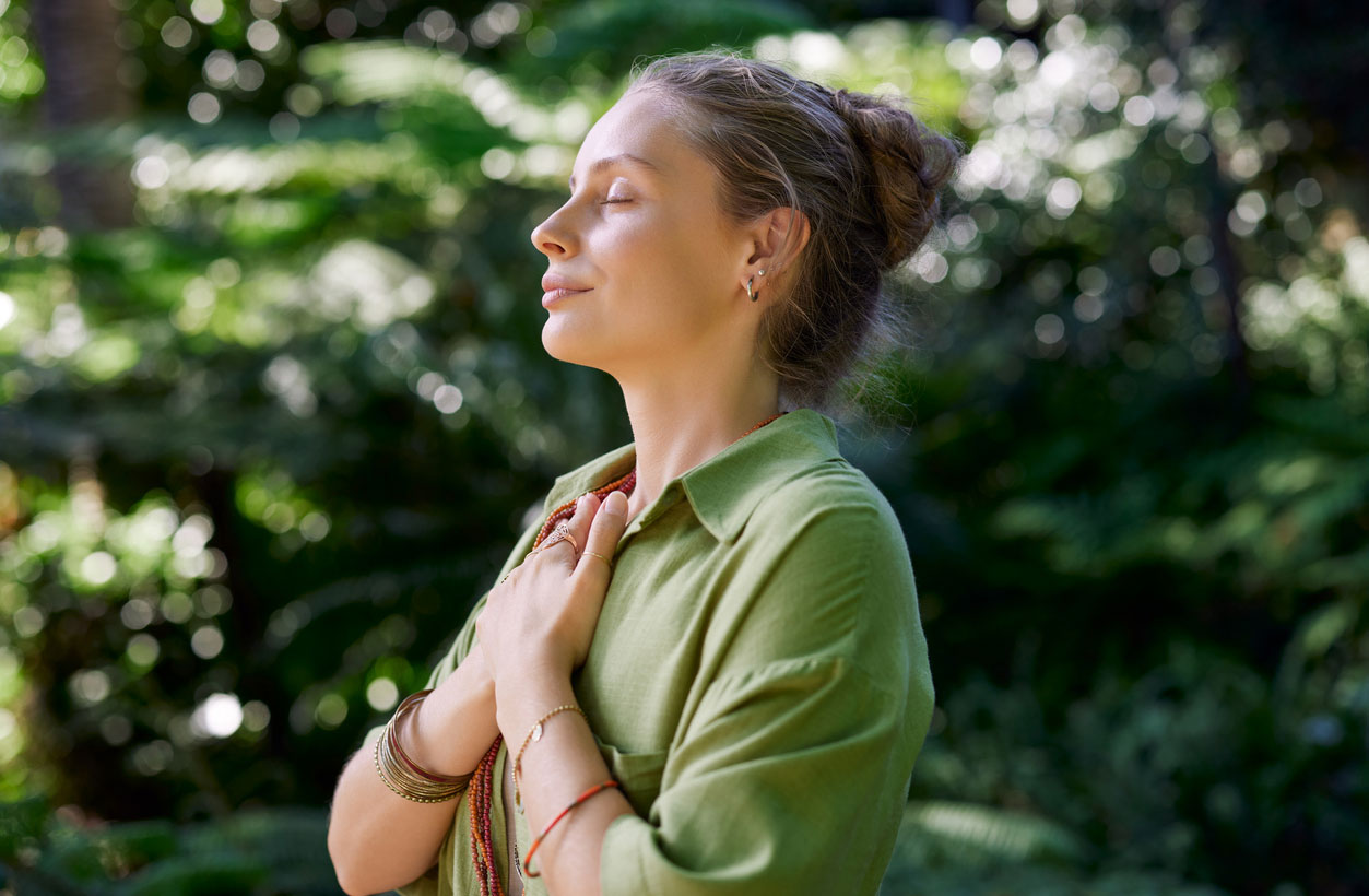 A peaceful woman practices stress management by placing her hands on her chest and breathing deeply, surrounded by nature.