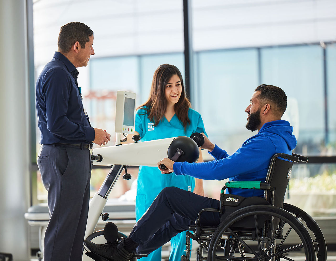 A man in a wheelchair uses an exercise machine, aided by two physical therapists.