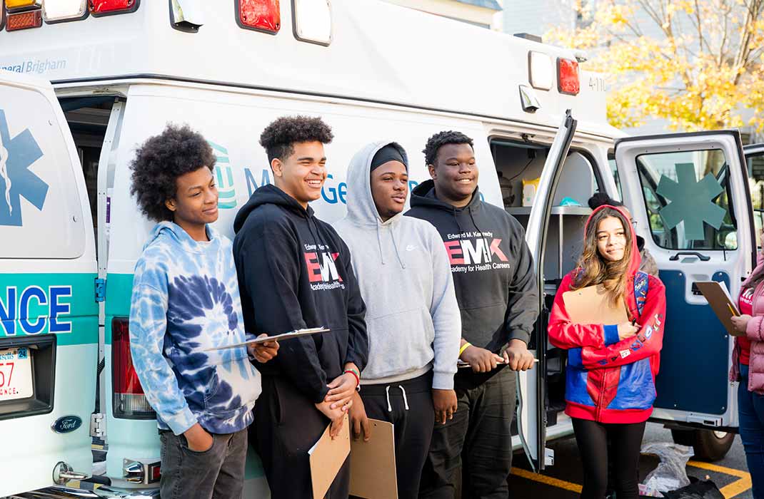 Five high school students smiling and standing in front of a Mass General Brigham ambulance