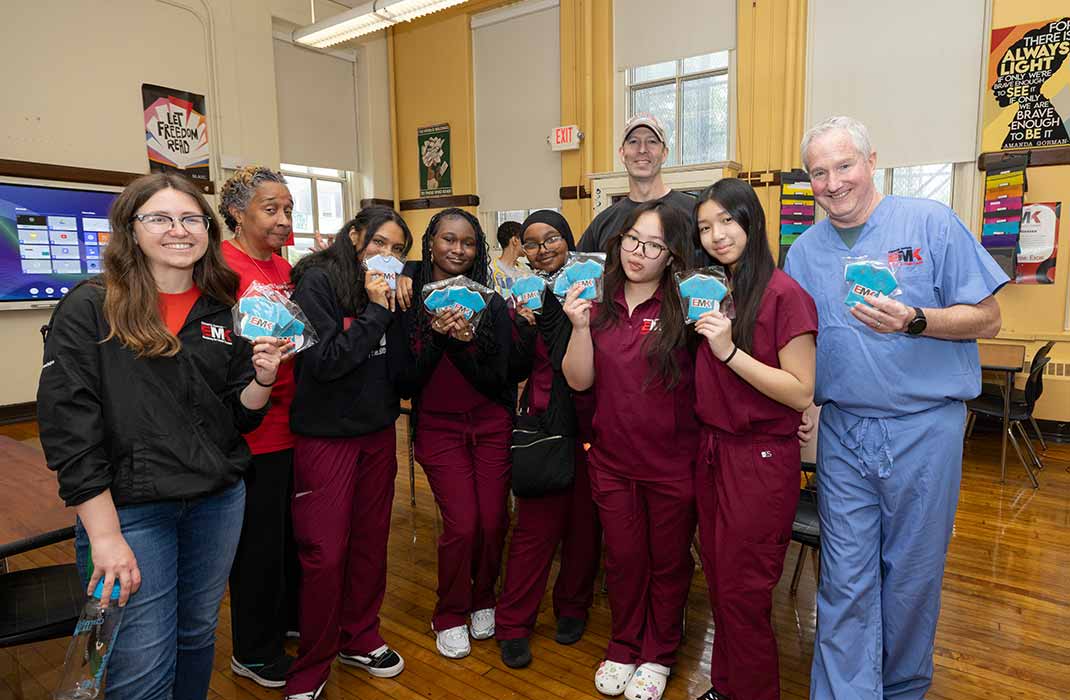 A group of nine people standing and smiling in a classroom. One of them is wearing blue scrubs.