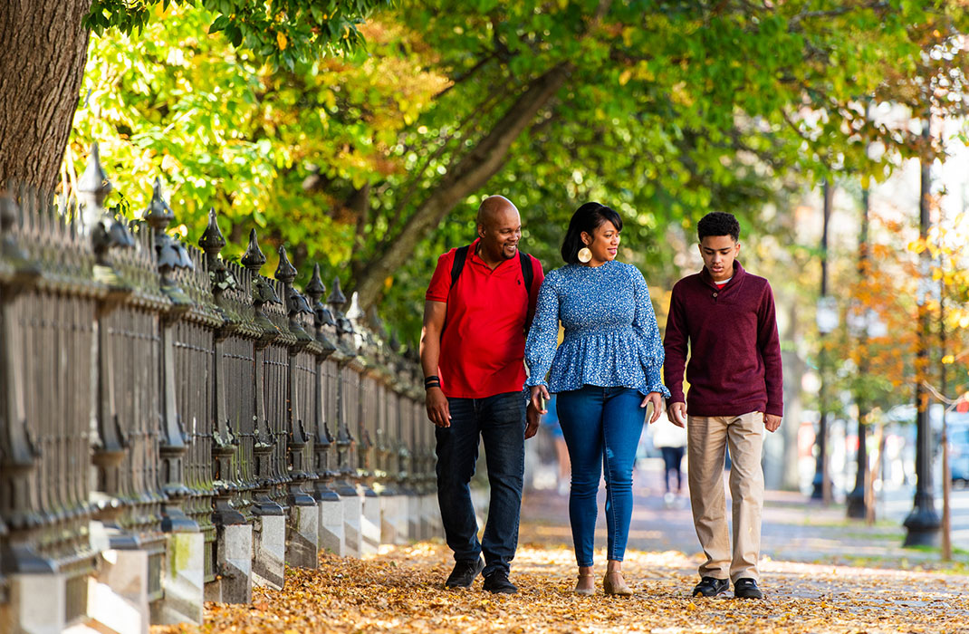 A mother, father, and teenaged son walk under the trees beside the railings at Boston Public Gardens.